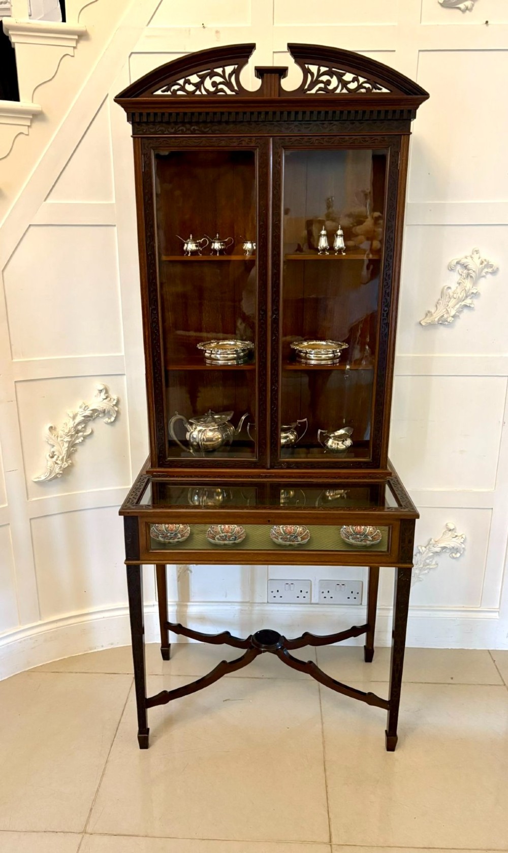 A carved mahogany bijouterie display cabinet featuring openwork fretwork pediment, glazed upper doors, and a glass-topped lower display compartment supported by slender legs with an X-shaped stretcher. View II