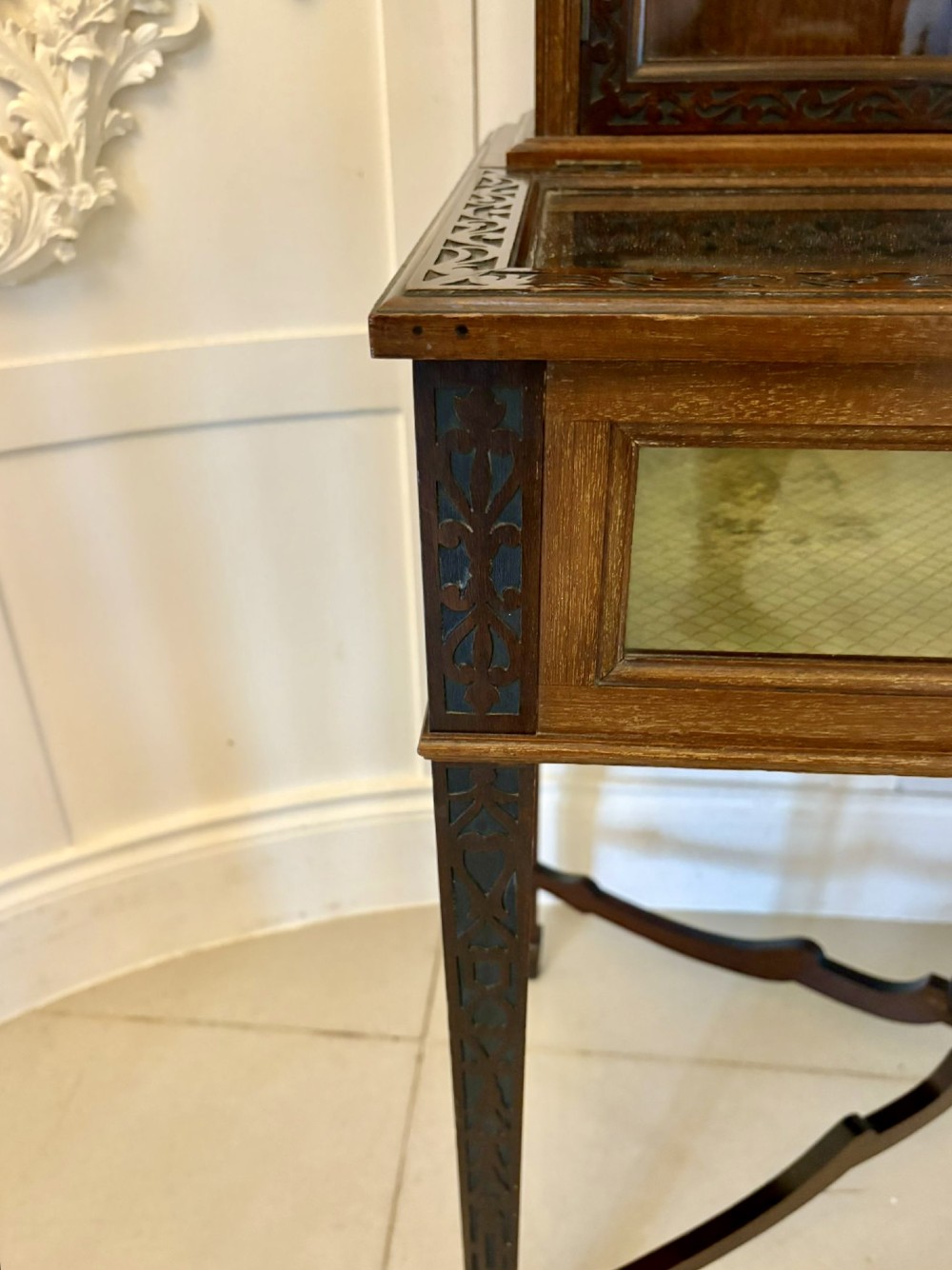 A carved mahogany bijouterie display cabinet featuring openwork fretwork pediment, glazed upper doors, and a glass-topped lower display compartment supported by slender legs with an X-shaped stretcher. View IV