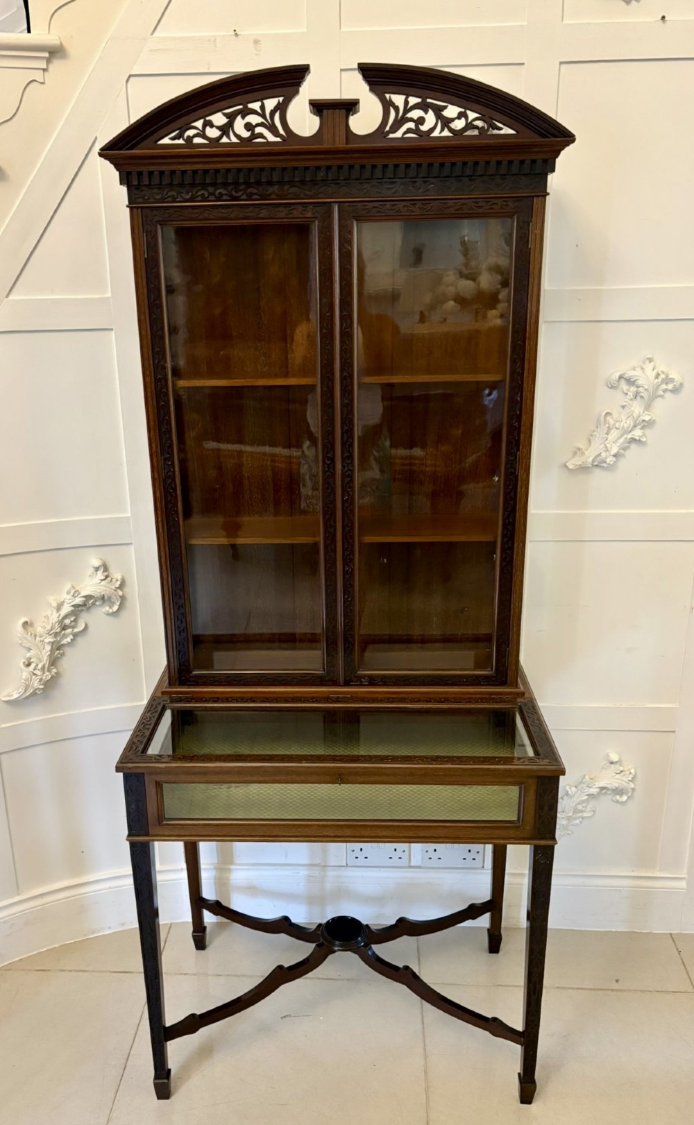 A carved mahogany bijouterie display cabinet featuring openwork fretwork pediment, glazed upper doors, and a glass-topped lower display compartment supported by slender legs with an X-shaped stretcher.