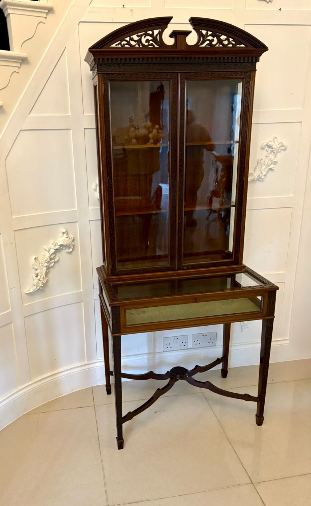 A carved mahogany bijouterie display cabinet featuring openwork fretwork pediment, glazed upper doors, and a glass-topped lower display compartment supported by slender legs with an X-shaped stretcher. View V