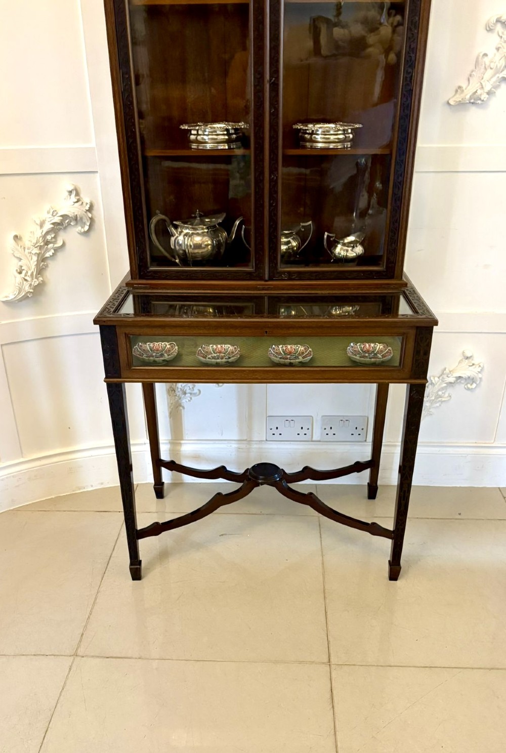 A carved mahogany bijouterie display cabinet featuring openwork fretwork pediment, glazed upper doors, and a glass-topped lower display compartment supported by slender legs with an X-shaped stretcher. View VI