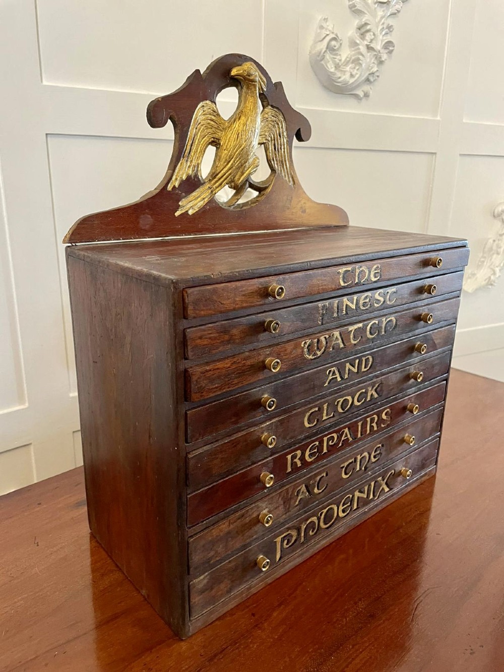 A dark wood Victorian desktop chest of eight shallow drawers featuring carved gilt lettering and a carved phoenix crest on the pediment. View IV