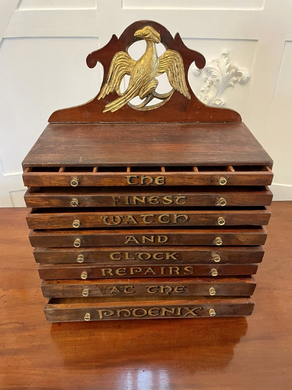 A dark wood Victorian desktop chest of eight shallow drawers featuring carved gilt lettering and a carved phoenix crest on the pediment. View V