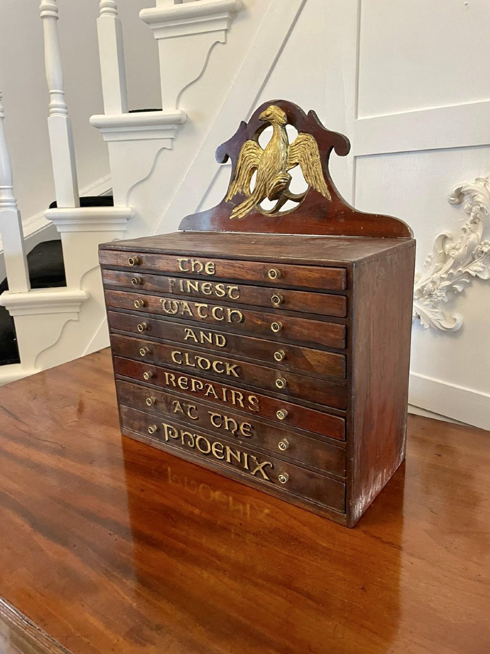 A dark wood Victorian desktop chest of eight shallow drawers featuring carved gilt lettering and a carved phoenix crest on the pediment. View VII