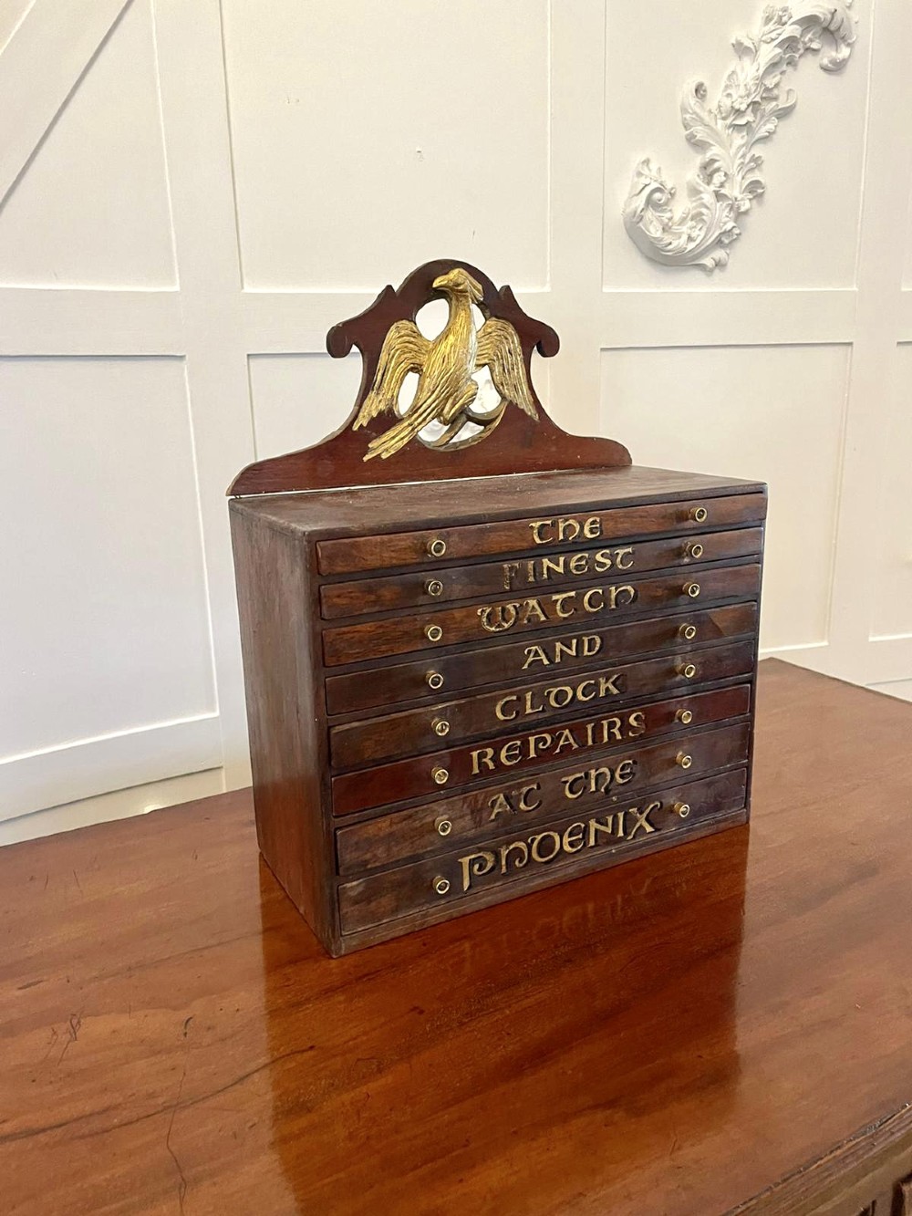A dark wood Victorian desktop chest of eight shallow drawers featuring carved gilt lettering and a carved phoenix crest on the pediment. View VIII