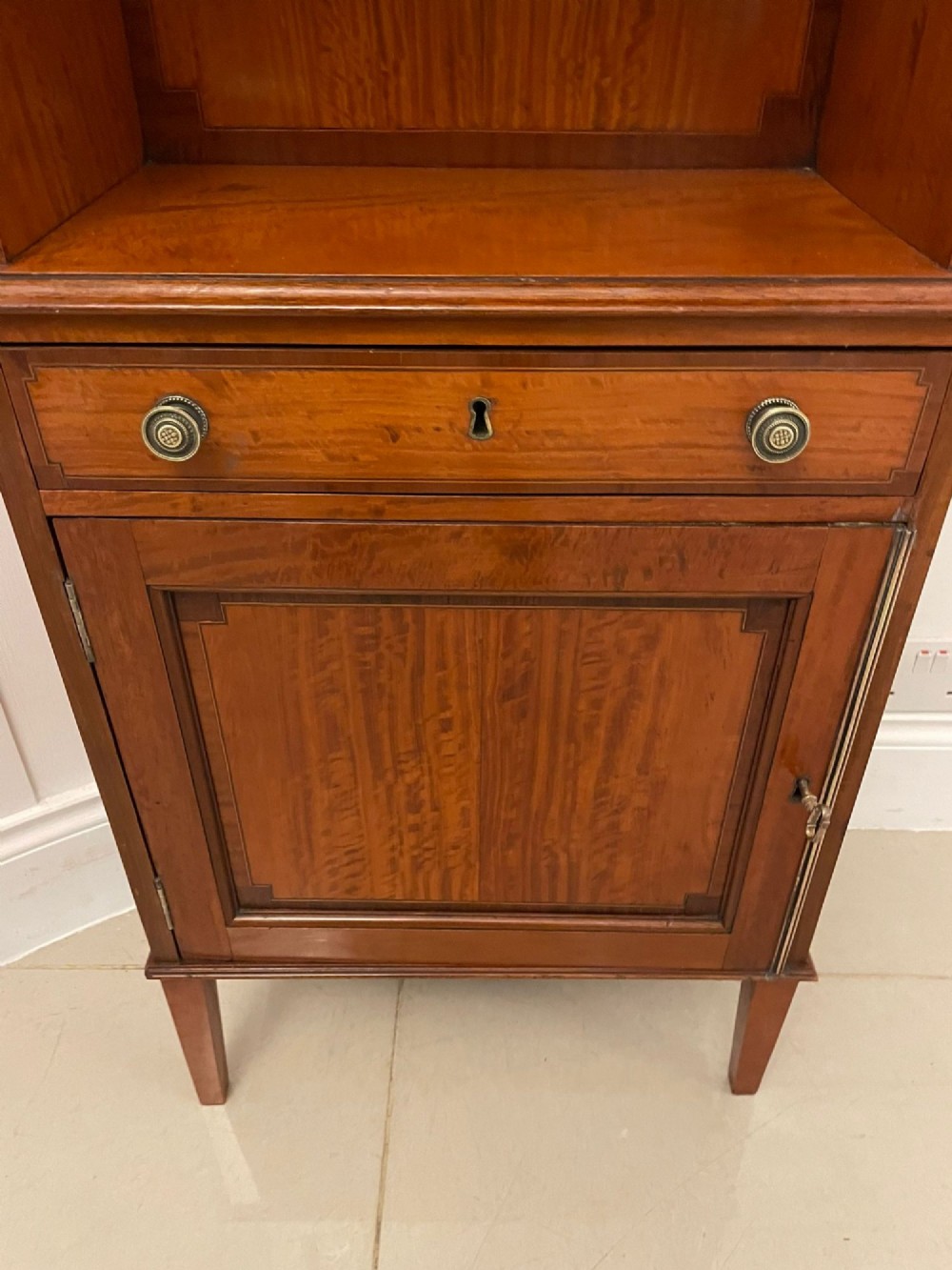 A pair of tall satinwood waterfall bookcases featuring tiered open shelves with brass gallery rails, single drawers, and lower panelled cupboards. View II
