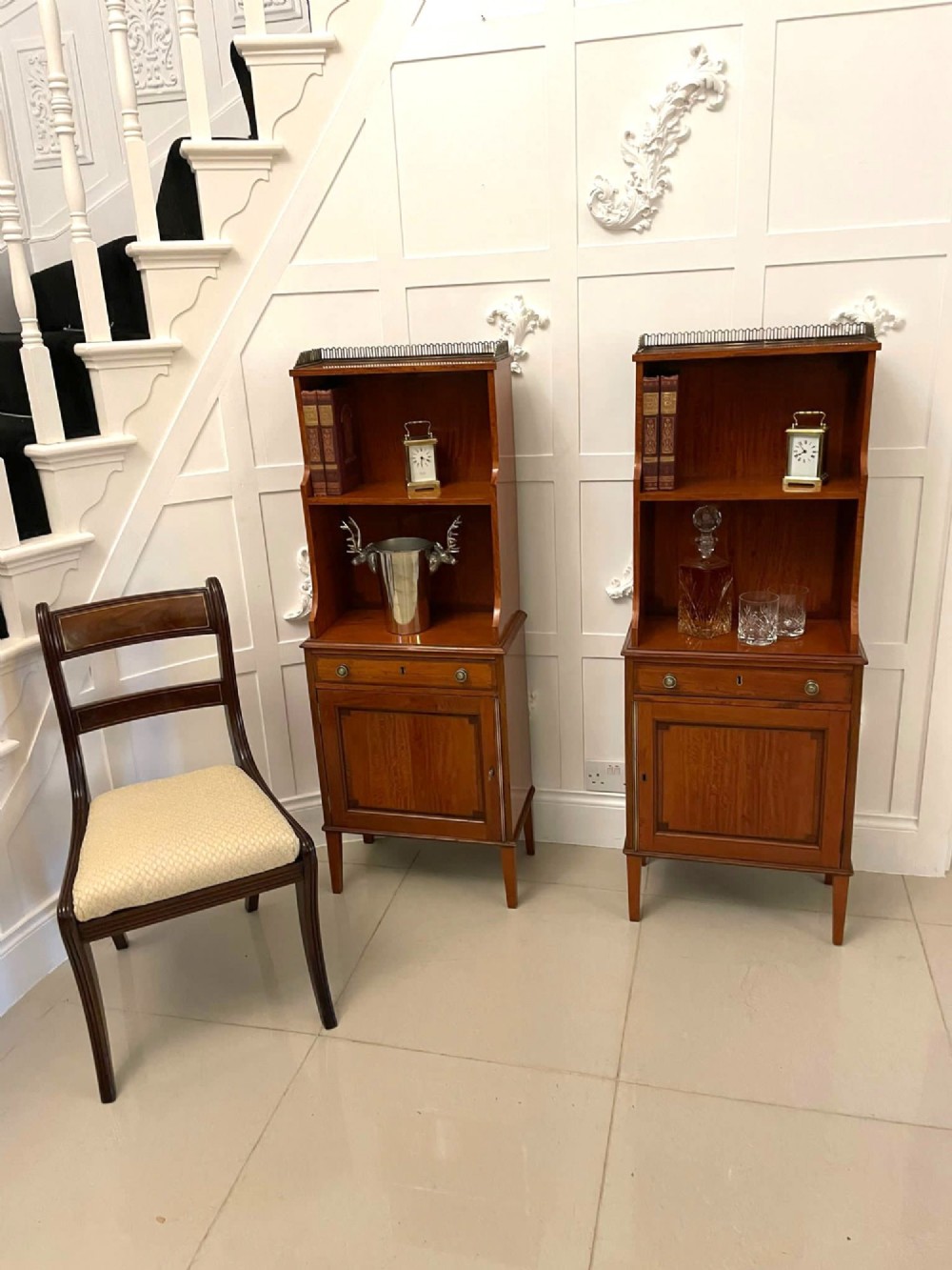 A pair of tall satinwood waterfall bookcases featuring tiered open shelves with brass gallery rails, single drawers, and lower panelled cupboards. View X