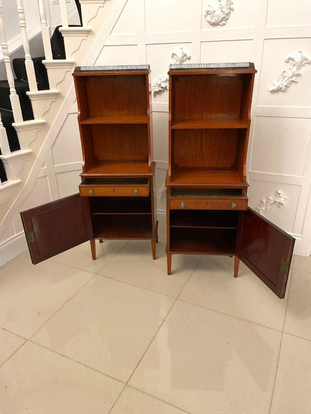 A pair of tall satinwood waterfall bookcases featuring tiered open shelves with brass gallery rails, single drawers, and lower panelled cupboards. View XIII