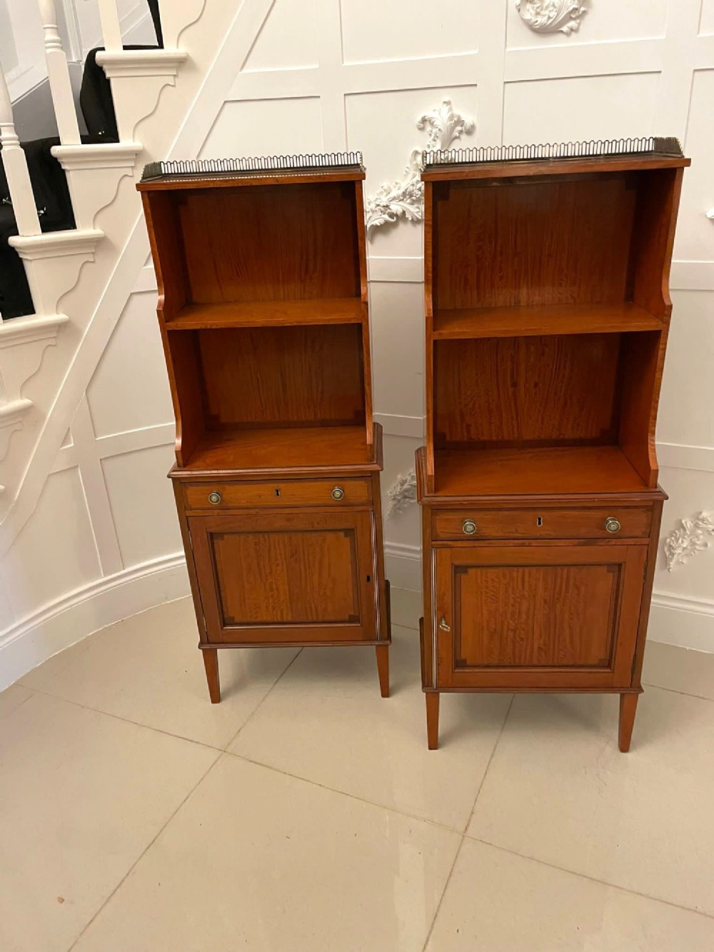 A pair of tall satinwood waterfall bookcases featuring tiered open shelves with brass gallery rails, single drawers, and lower panelled cupboards. View XIV