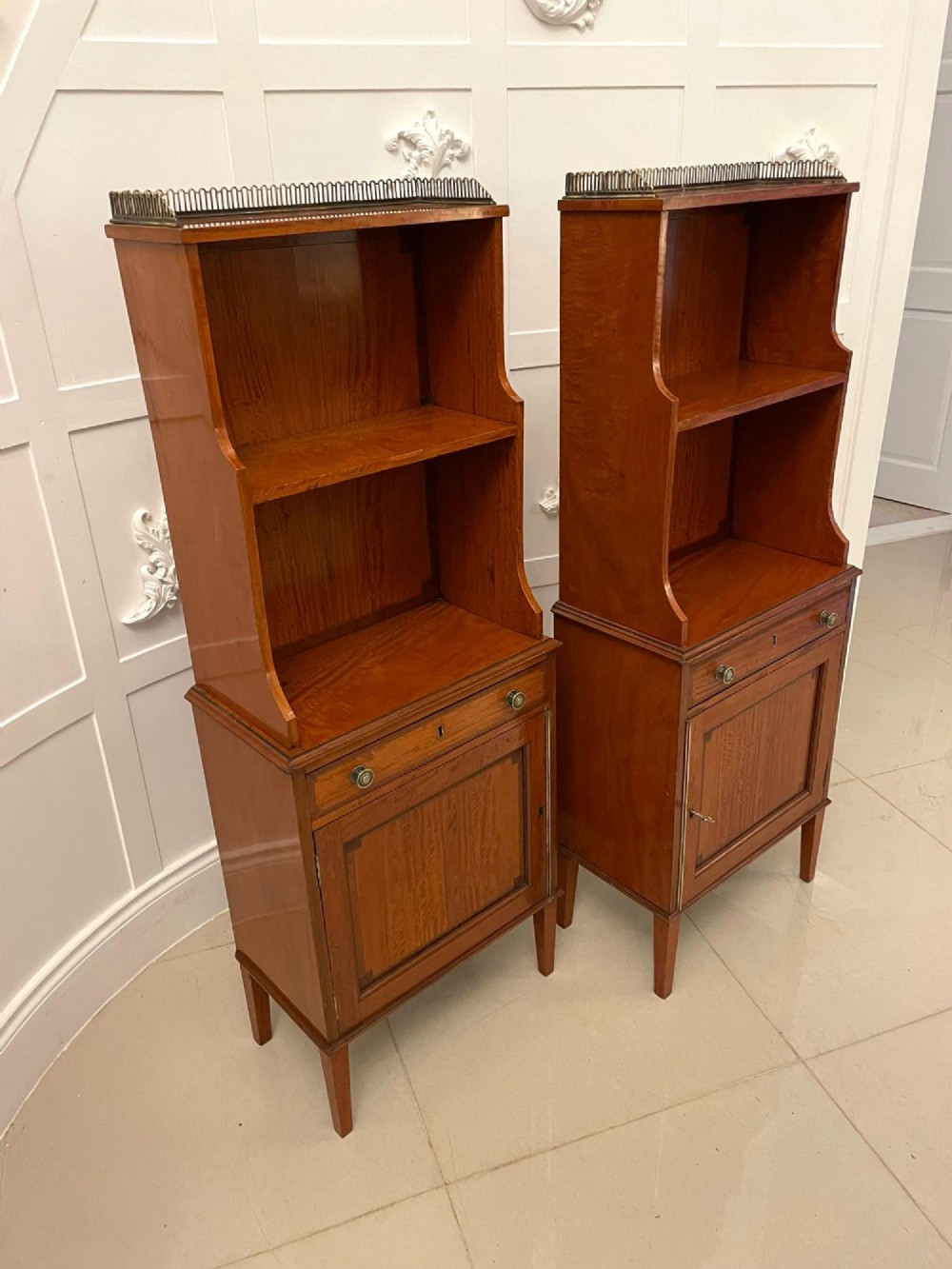 A pair of tall satinwood waterfall bookcases featuring tiered open shelves with brass gallery rails, single drawers, and lower panelled cupboards. View XV