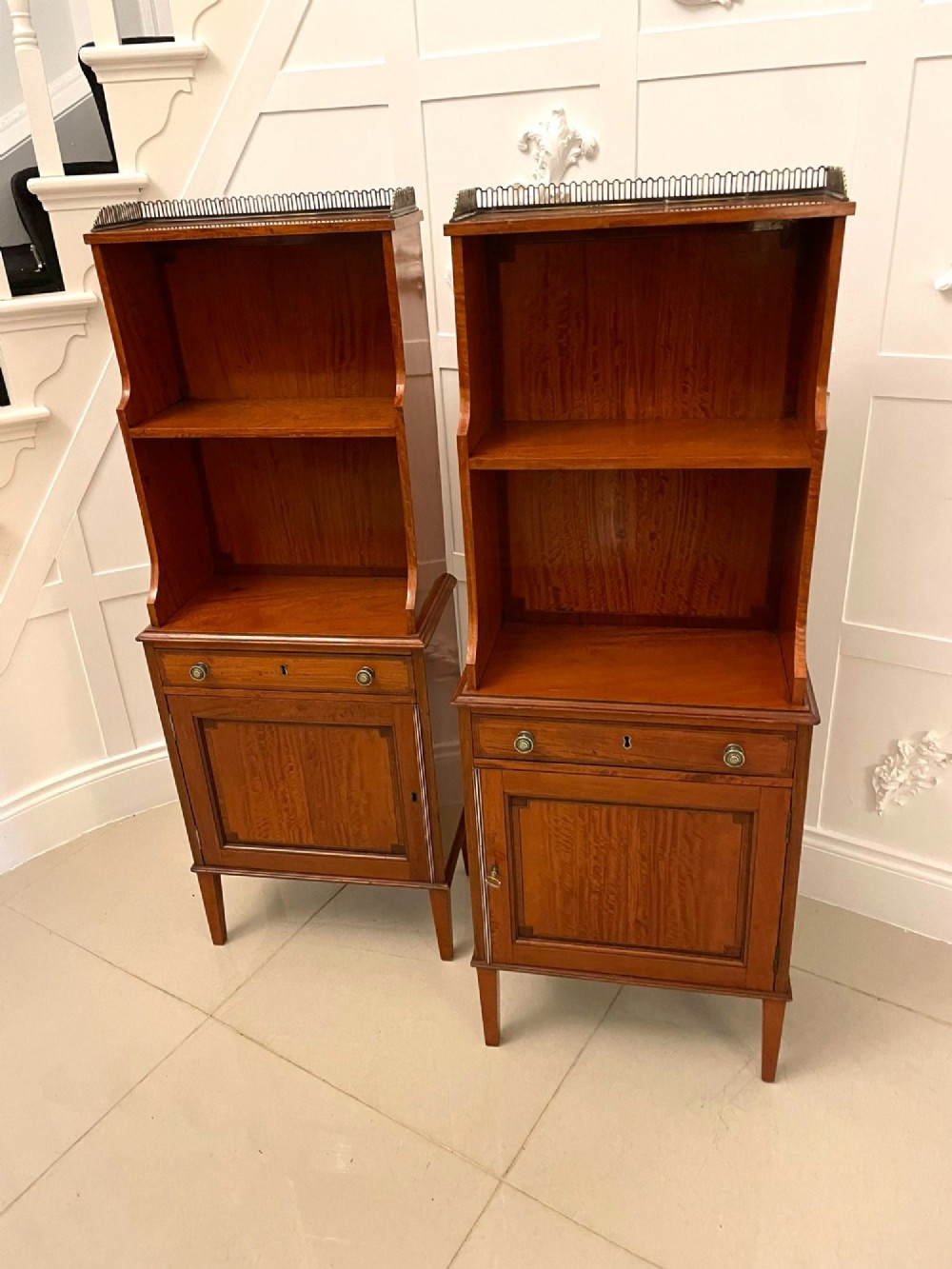 A pair of tall satinwood waterfall bookcases featuring tiered open shelves with brass gallery rails, single drawers, and lower panelled cupboards.