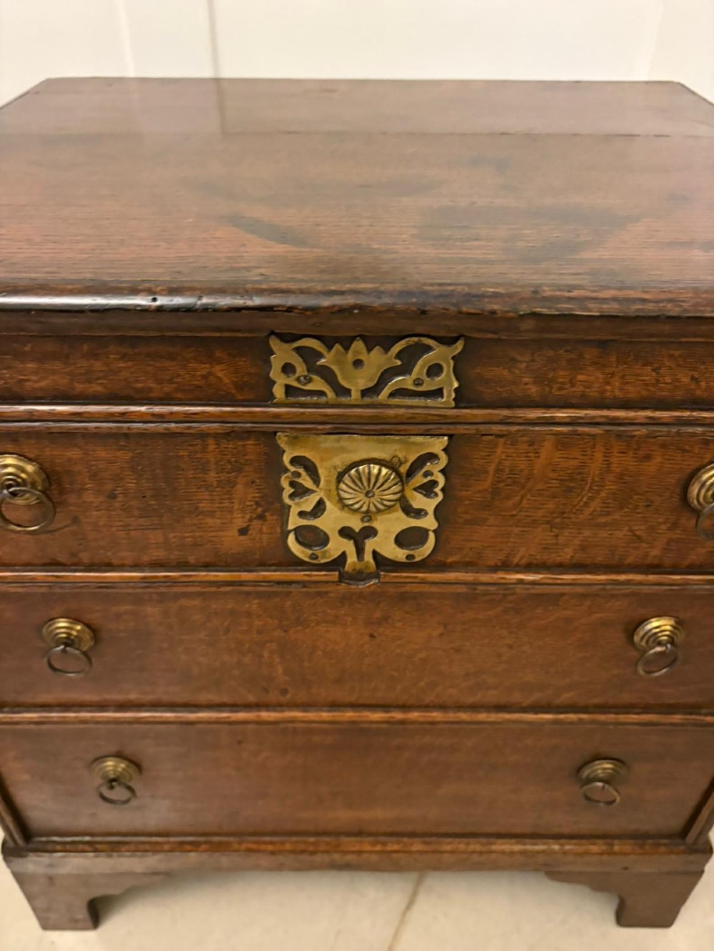 An early 18th-century dark oak box coffer styled as a miniature chest of drawers with decorative brass escutcheons, ring handles, and bracket feet. View X