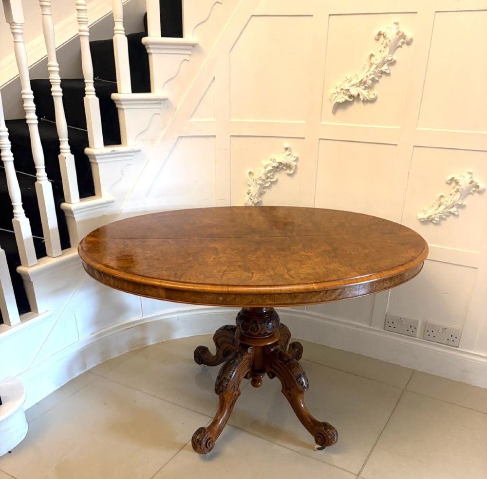 An antique Victorian oval tilt-top table made of burr walnut with a four-star carved pedestal base and white ceramic castors, shown with the tabletop tilted vertically. View IX