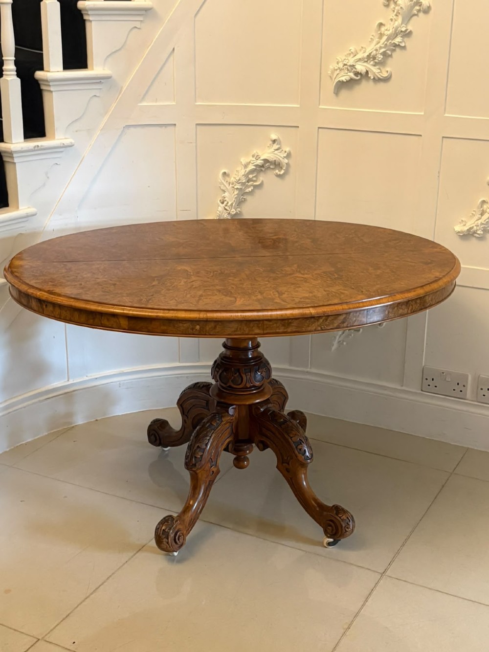 An antique Victorian oval tilt-top table made of burr walnut with a four-star carved pedestal base and white ceramic castors, shown with the tabletop tilted vertically. View XI