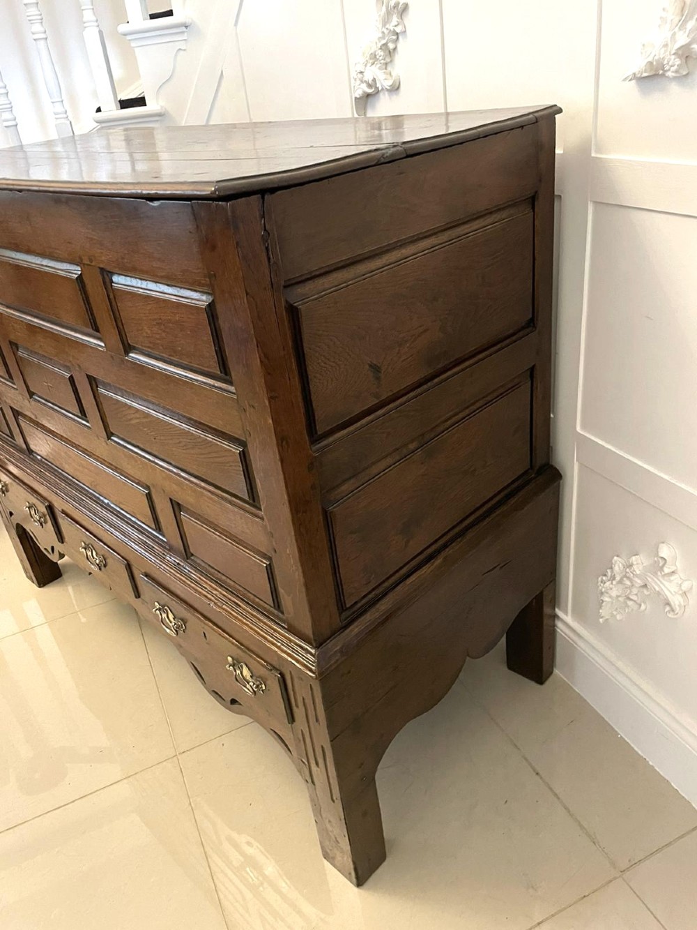 An 18th-century oak coffer elevated on a stand, with geometric panel moulding across the front lid section, lower brass-handled drawers, and square legs. View XII