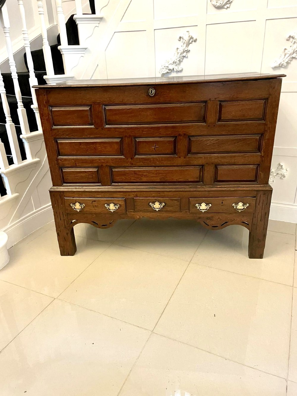 An 18th-century oak coffer elevated on a stand, with geometric panel moulding across the front lid section, lower brass-handled drawers, and square legs.