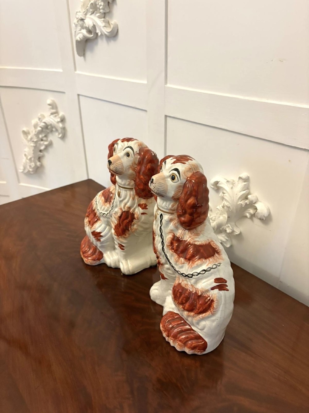 A pair of Victorian Staffordshire ceramic spaniel figurines with painted russet spots and black chain collars seated on a dark wooden surface. View X