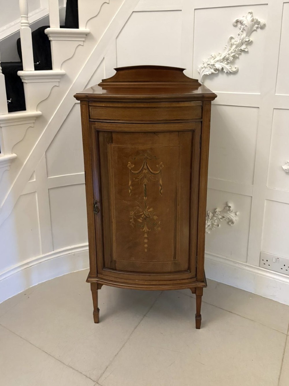An Edwardian mahogany bow-fronted side cabinet featuring ribbon and urn marquetry inlay, a raised gallery back, and tapered square legs.