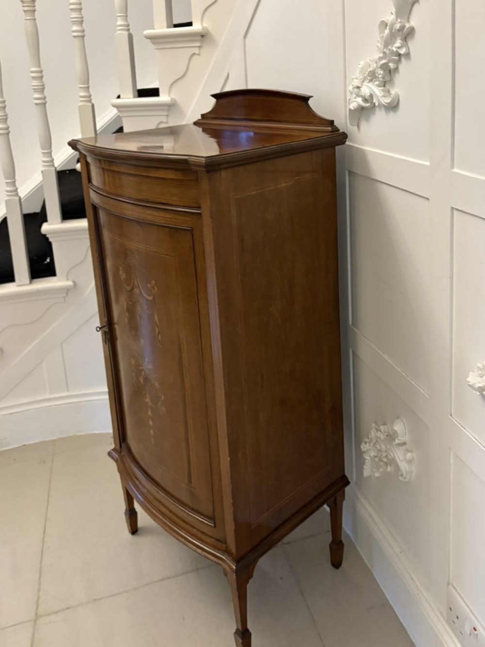 An Edwardian mahogany bow-fronted side cabinet featuring ribbon and urn marquetry inlay, a raised gallery back, and tapered square legs. View XI