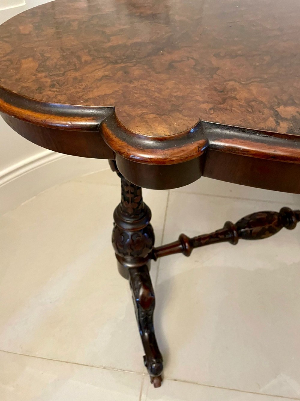 An antique Victorian oval centre table featuring a figured burr walnut top with moulded edges, carved end supports, a turned stretcher, and brass castors. View V