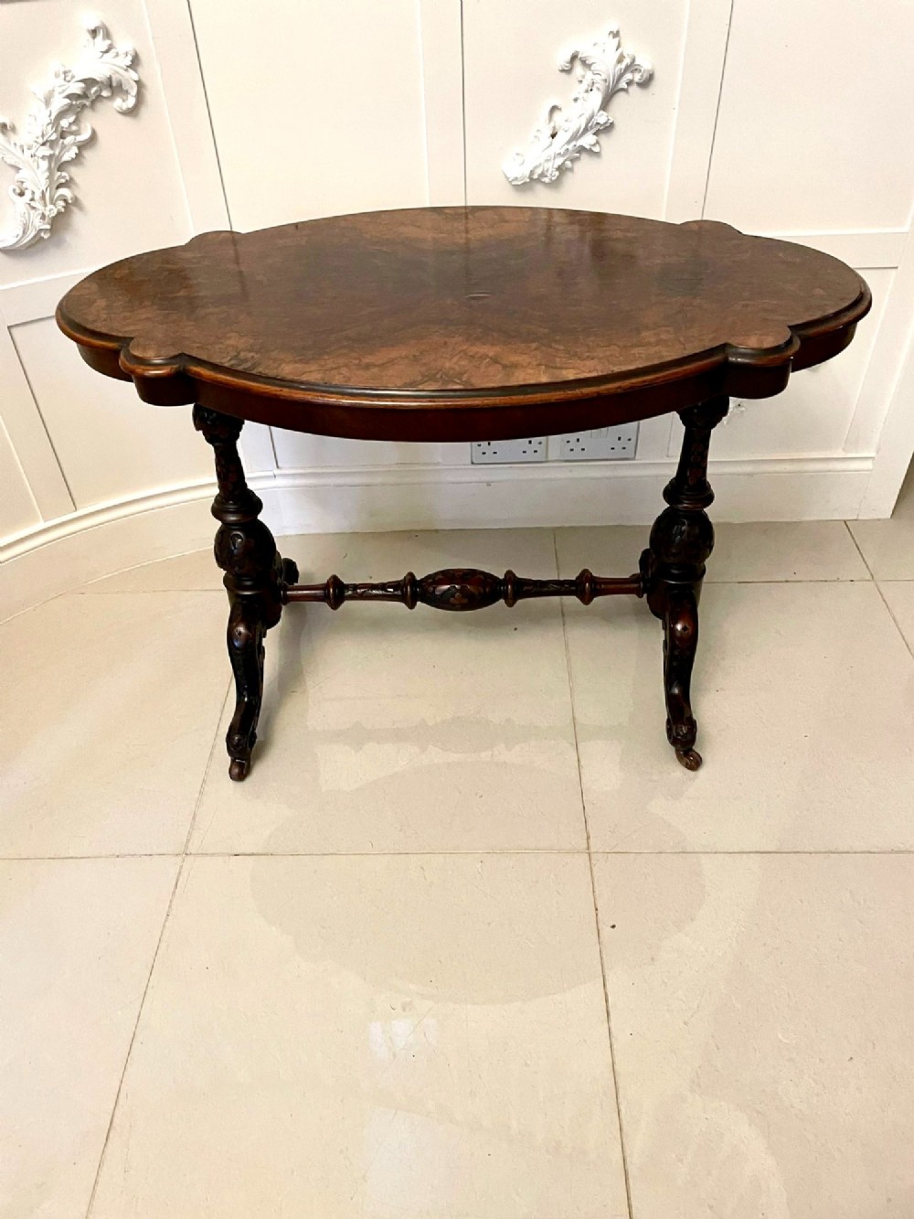 An antique Victorian oval centre table featuring a figured burr walnut top with moulded edges, carved end supports, a turned stretcher, and brass castors. View VII