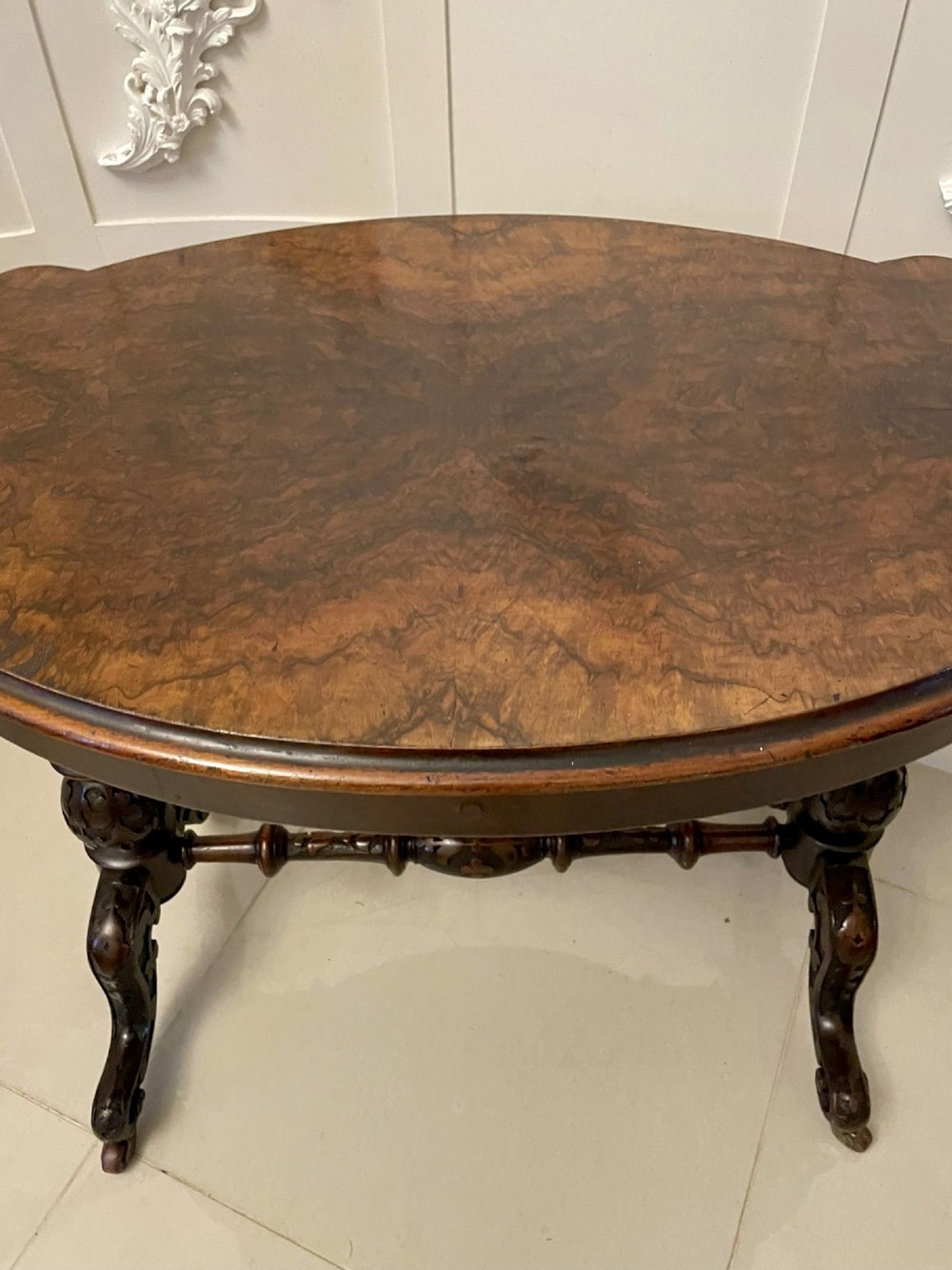 An antique Victorian oval centre table featuring a figured burr walnut top with moulded edges, carved end supports, a turned stretcher, and brass castors. View VIII