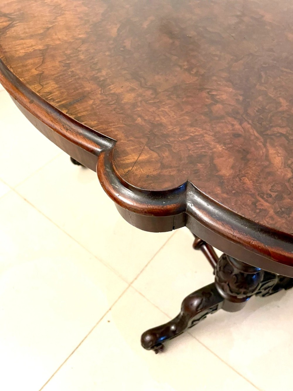 An antique Victorian oval centre table featuring a figured burr walnut top with moulded edges, carved end supports, a turned stretcher, and brass castors. View IX