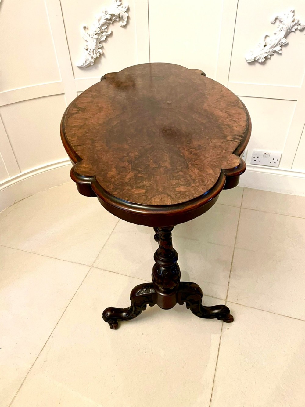 An antique Victorian oval centre table featuring a figured burr walnut top with moulded edges, carved end supports, a turned stretcher, and brass castors. View XII