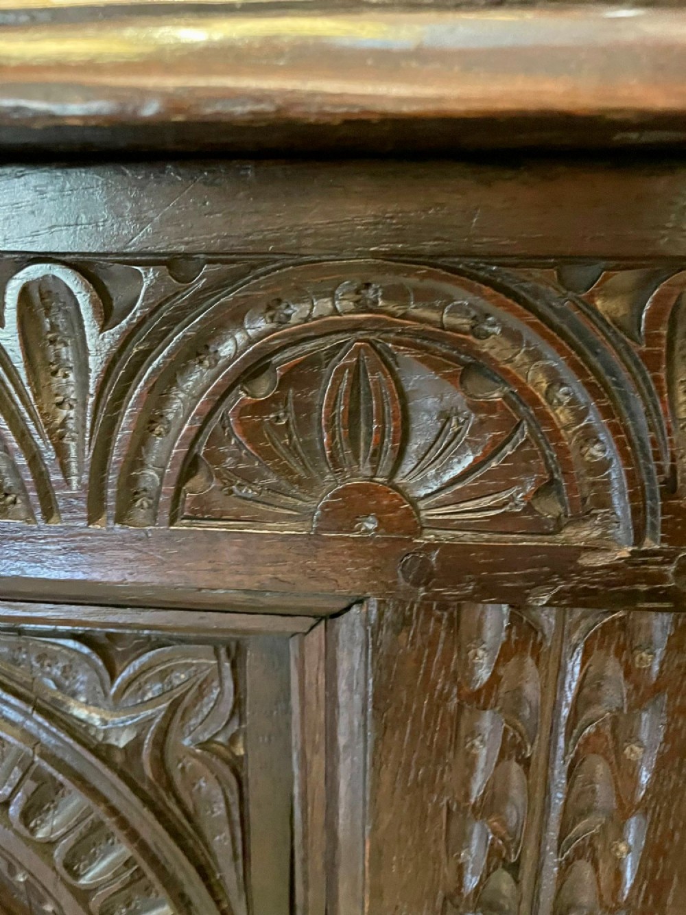 A dark carved oak coffer featuring three arcaded front panels, carved border details, top iron hinges, and turned bun feet. View V