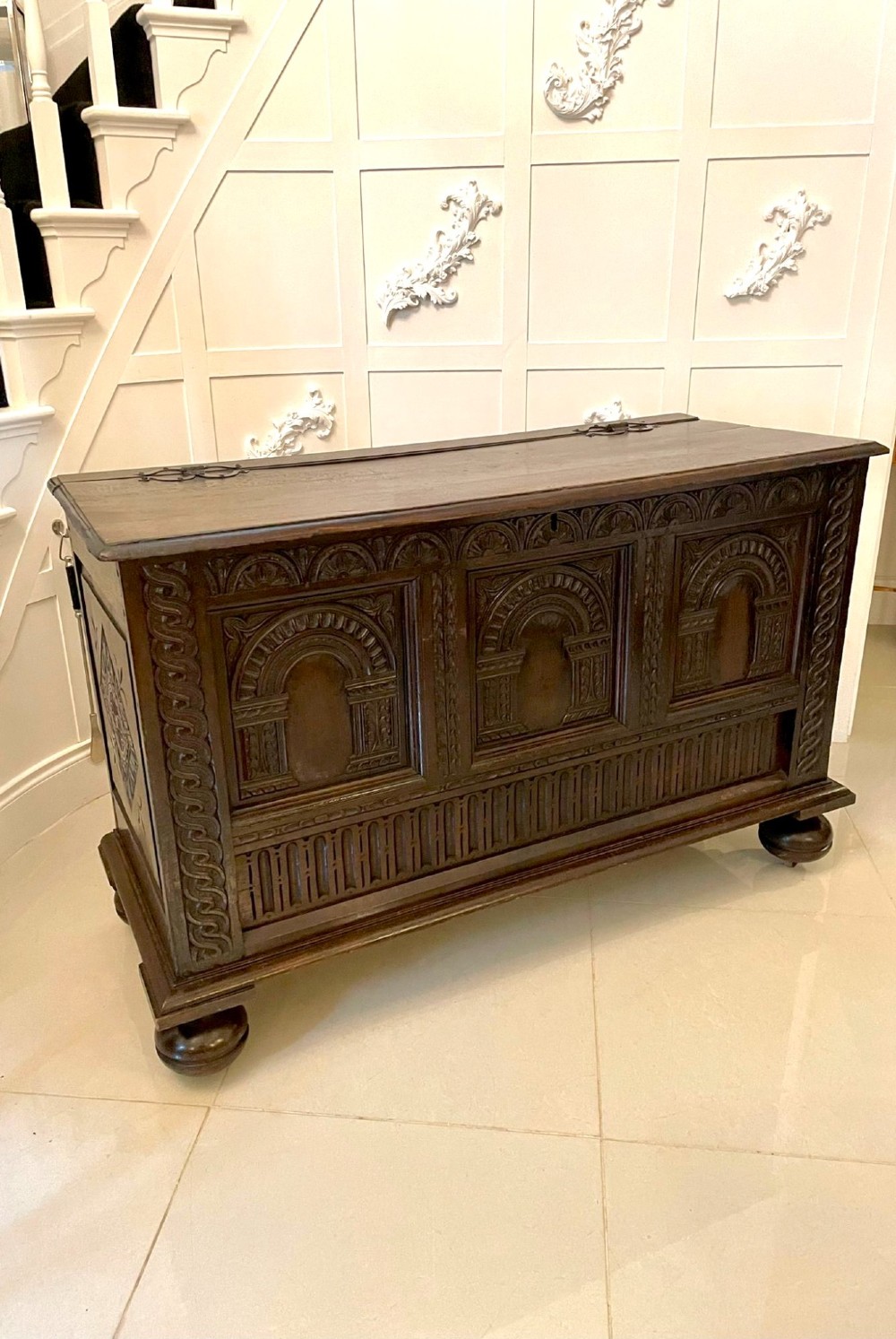 A dark carved oak coffer featuring three arcaded front panels, carved border details, top iron hinges, and turned bun feet.