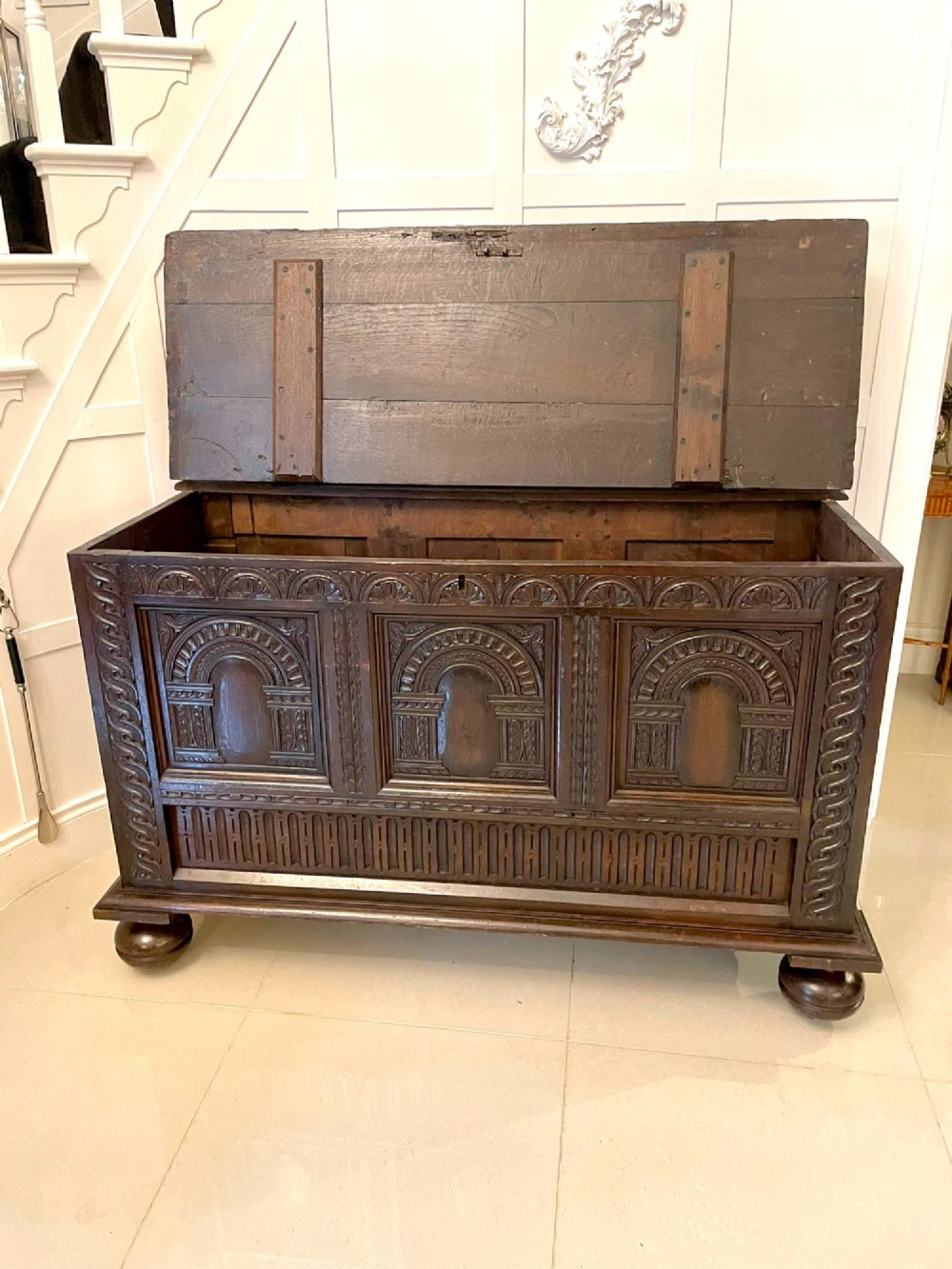 A dark carved oak coffer featuring three arcaded front panels, carved border details, top iron hinges, and turned bun feet. View XIV