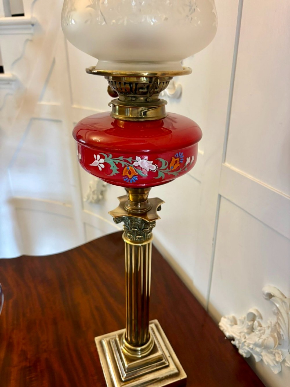 An antique Victorian oil lamp featuring a stepped square brass base, a fluted Corinthian column stem, an enamel-painted red glass font, and an etched cranberry glass frilled shade. View II