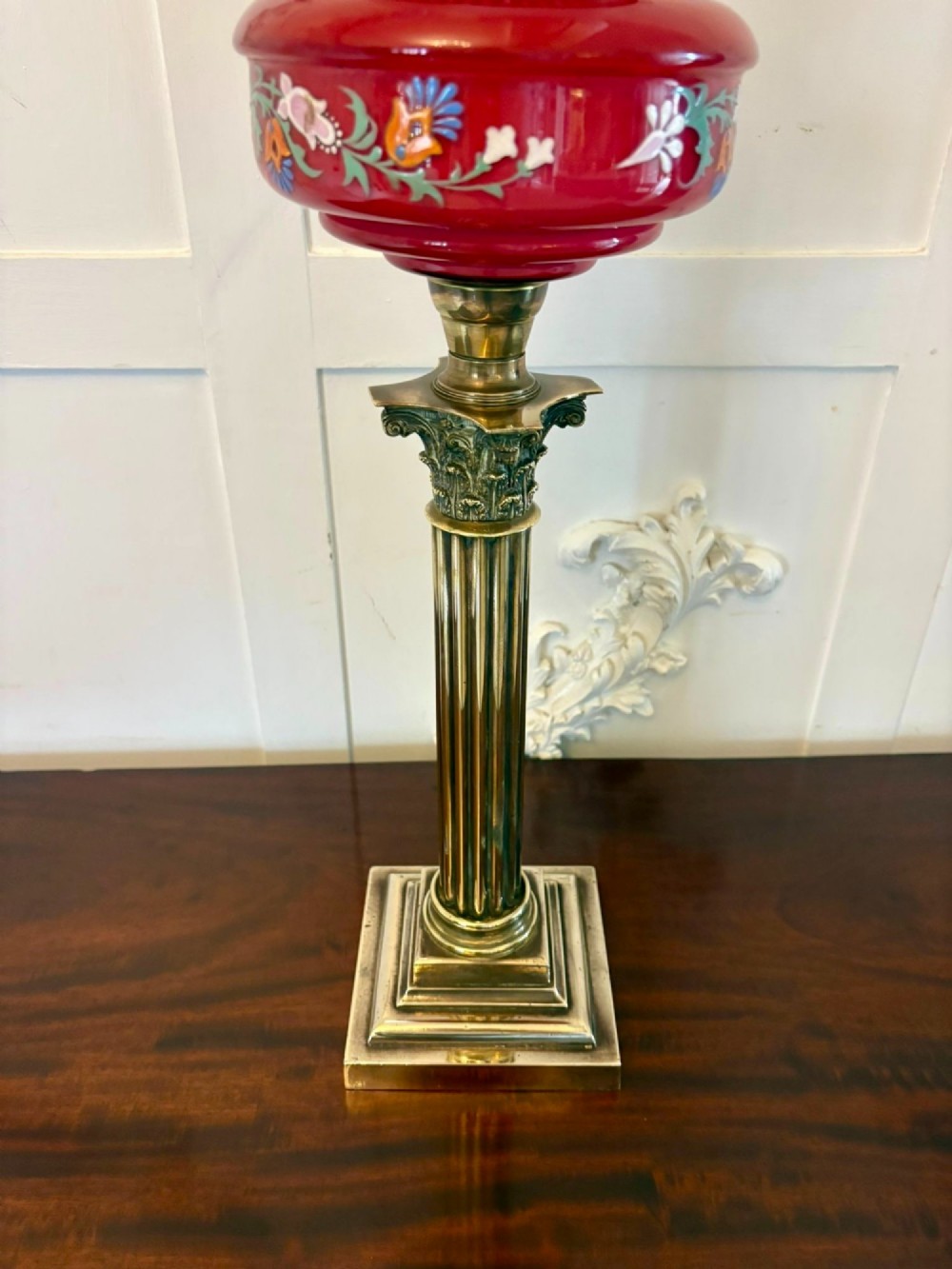 An antique Victorian oil lamp featuring a stepped square brass base, a fluted Corinthian column stem, an enamel-painted red glass font, and an etched cranberry glass frilled shade. View IV