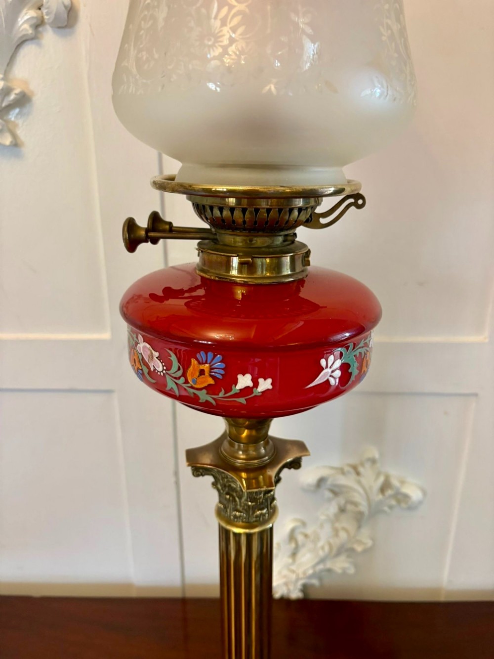 An antique Victorian oil lamp featuring a stepped square brass base, a fluted Corinthian column stem, an enamel-painted red glass font, and an etched cranberry glass frilled shade. View VI