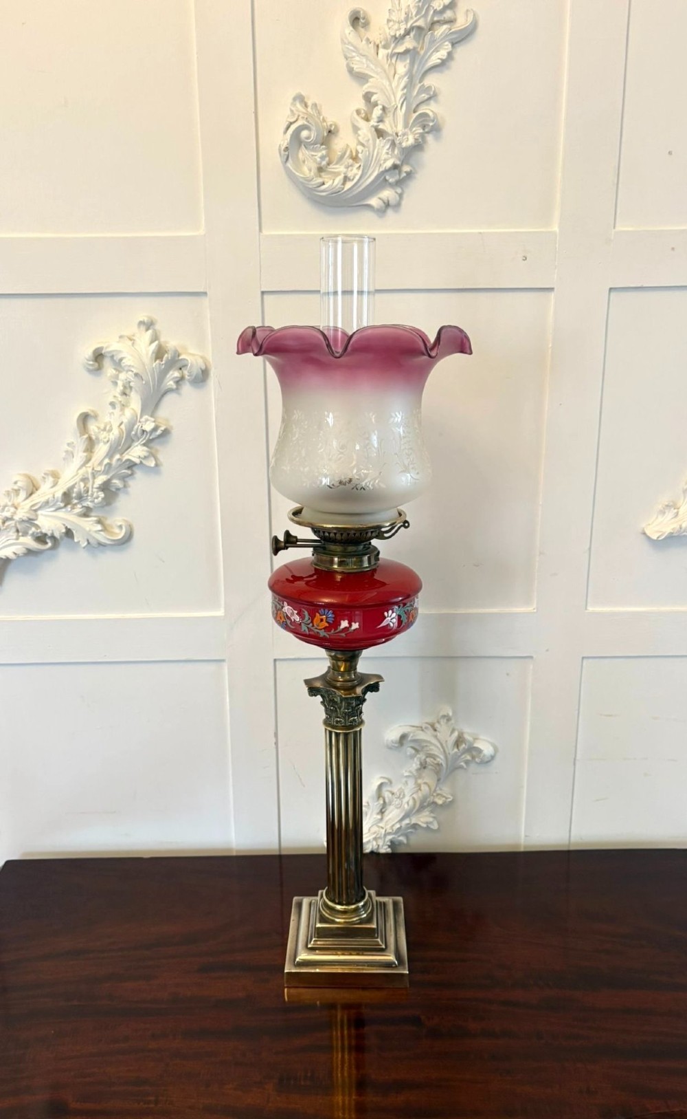 An antique Victorian oil lamp featuring a stepped square brass base, a fluted Corinthian column stem, an enamel-painted red glass font, and an etched cranberry glass frilled shade.