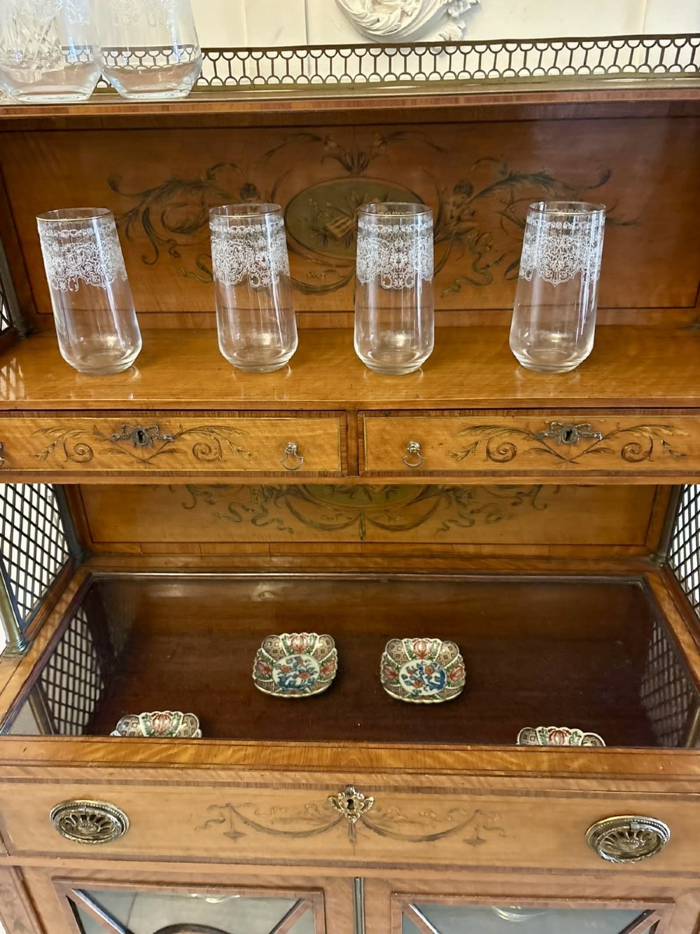 A Georgian Sheraton style satinwood display cabinet featuring upper open shelves with brass grilles and marquetry panels, positioned above a lower glazed twin-door cabinet with oval astragal bars. View V