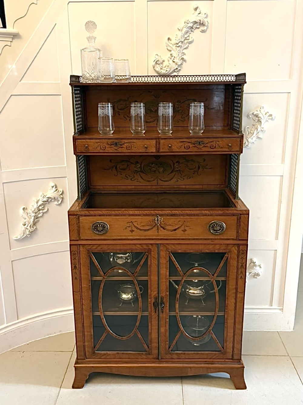 A Georgian Sheraton style satinwood display cabinet featuring upper open shelves with brass grilles and marquetry panels, positioned above a lower glazed twin-door cabinet with oval astragal bars. View VI