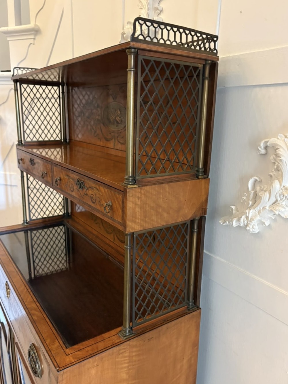 A Georgian Sheraton style satinwood display cabinet featuring upper open shelves with brass grilles and marquetry panels, positioned above a lower glazed twin-door cabinet with oval astragal bars. View VII