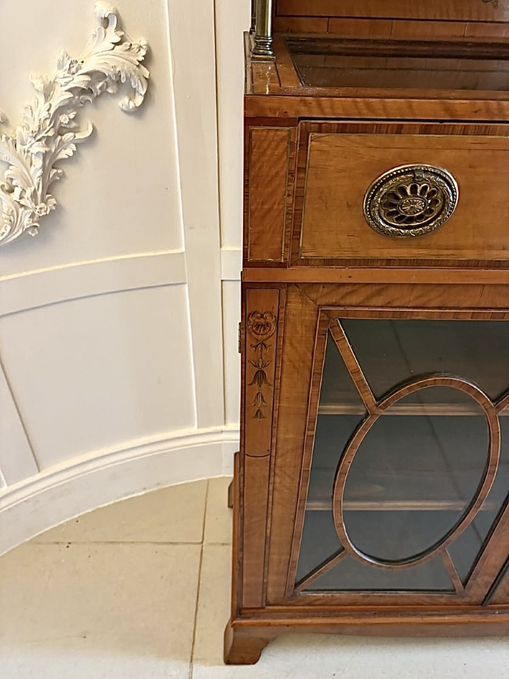 A Georgian Sheraton style satinwood display cabinet featuring upper open shelves with brass grilles and marquetry panels, positioned above a lower glazed twin-door cabinet with oval astragal bars. View VIII