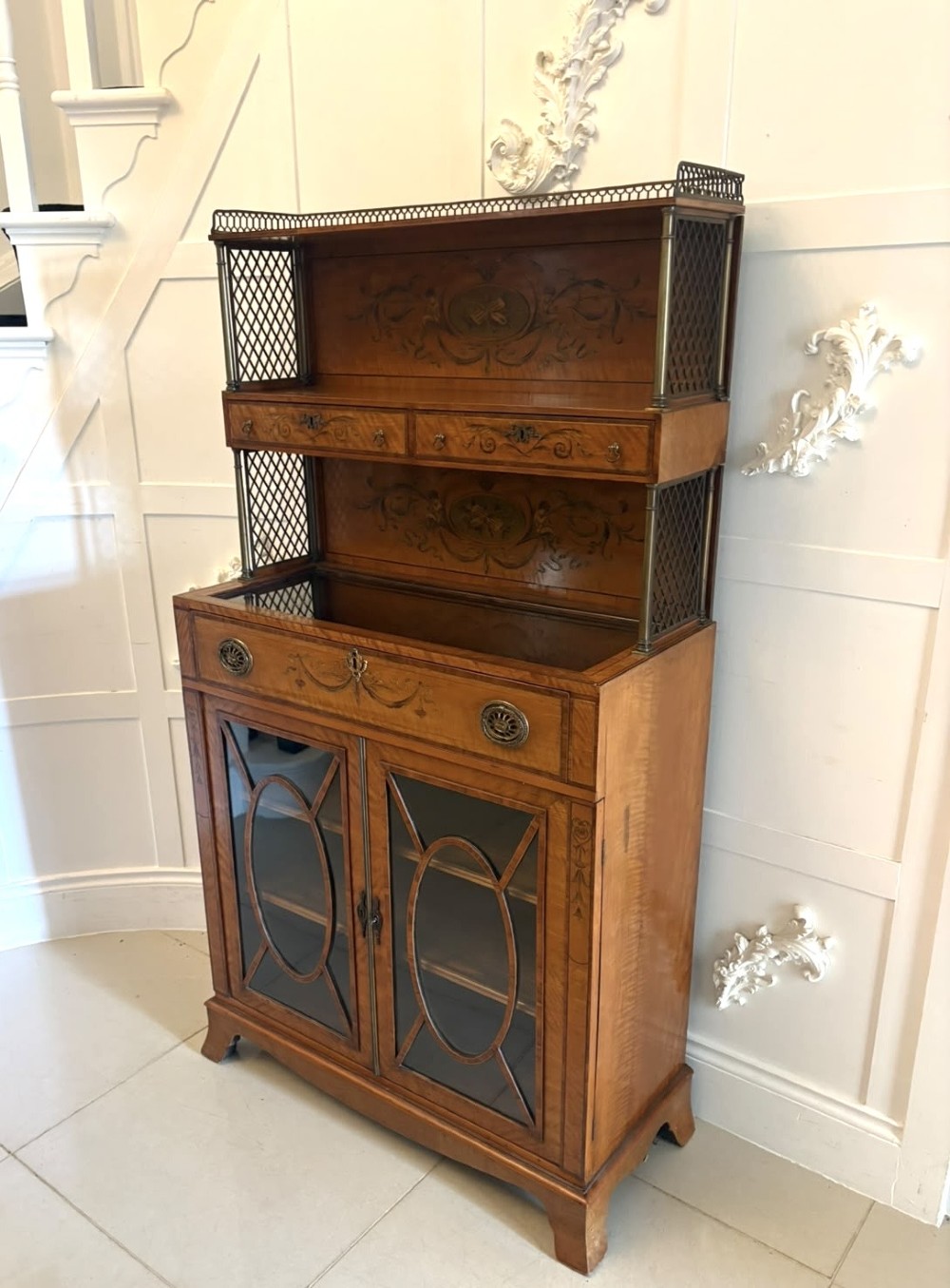 A Georgian Sheraton style satinwood display cabinet featuring upper open shelves with brass grilles and marquetry panels, positioned above a lower glazed twin-door cabinet with oval astragal bars. View XIV