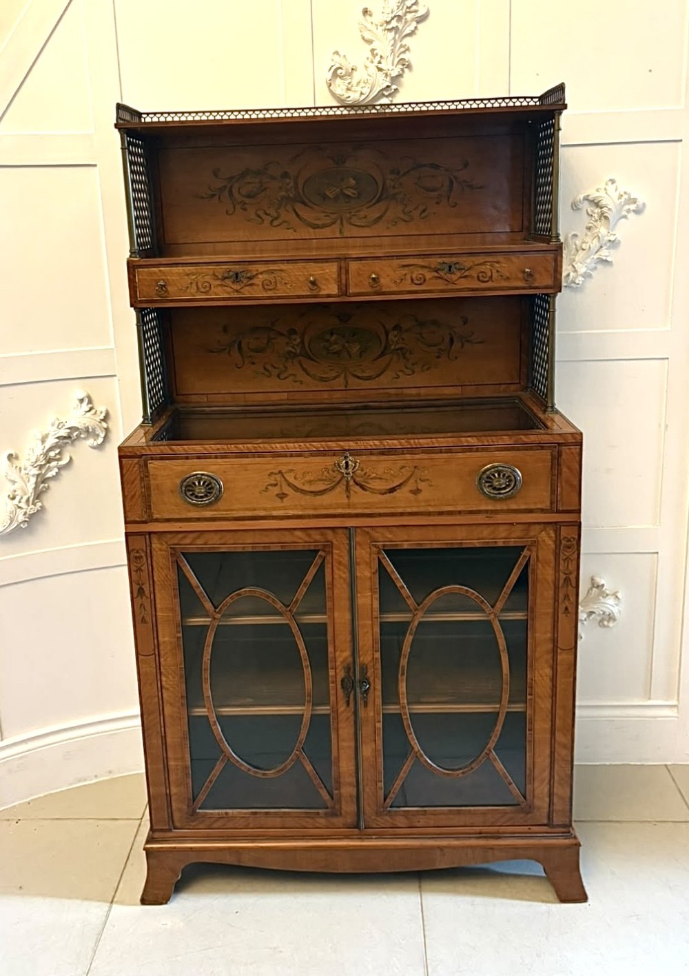 A Georgian Sheraton style satinwood display cabinet featuring upper open shelves with brass grilles and marquetry panels, positioned above a lower glazed twin-door cabinet with oval astragal bars. View XV
