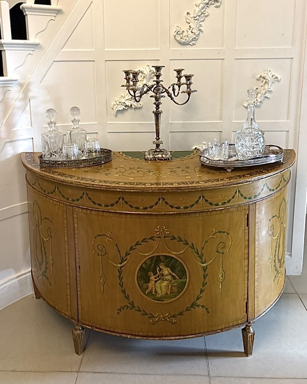 A semicircular satinwood demi-lune cabinet featuring hand-painted neoclassical leaf swags, a central figural medallion, and short tapered feet. View V