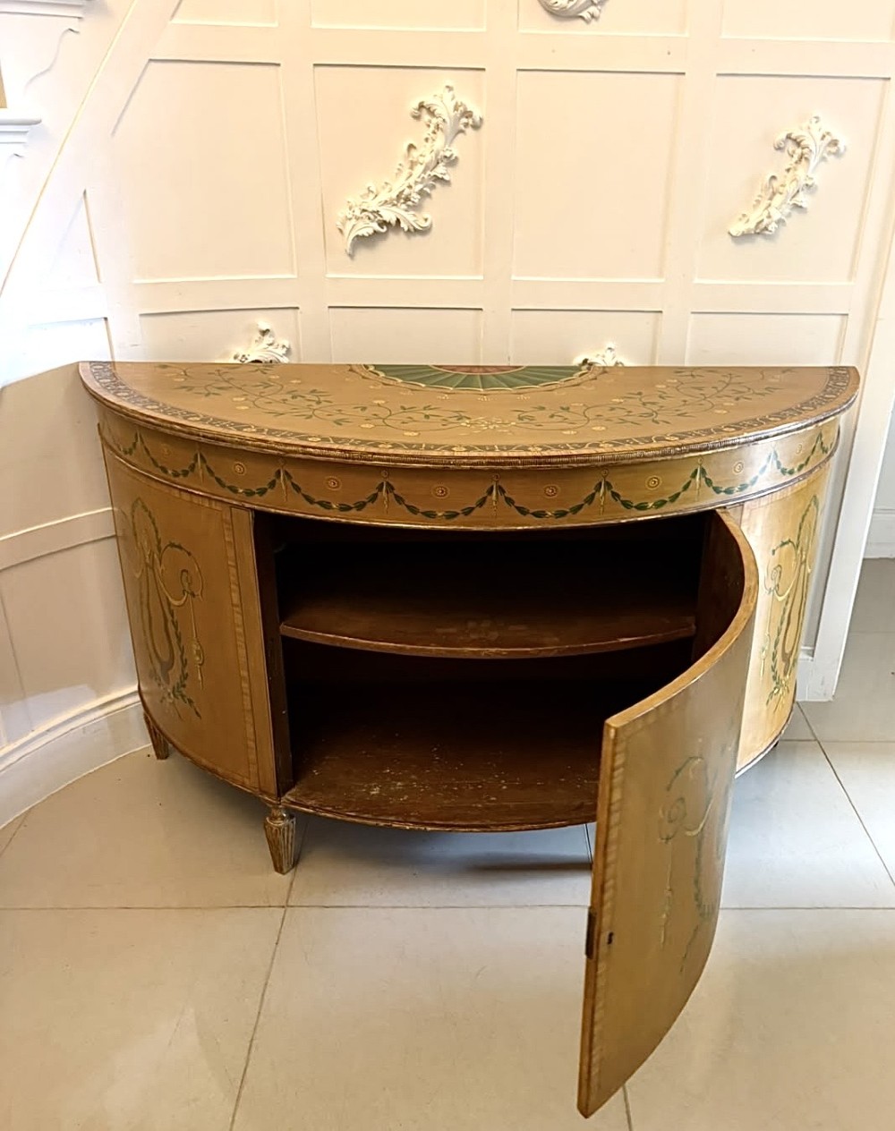 A semicircular satinwood demi-lune cabinet featuring hand-painted neoclassical leaf swags, a central figural medallion, and short tapered feet. View X