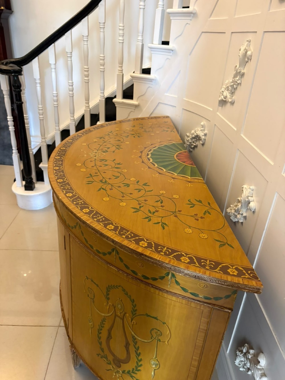 A semicircular satinwood demi-lune cabinet featuring hand-painted neoclassical leaf swags, a central figural medallion, and short tapered feet. View XII