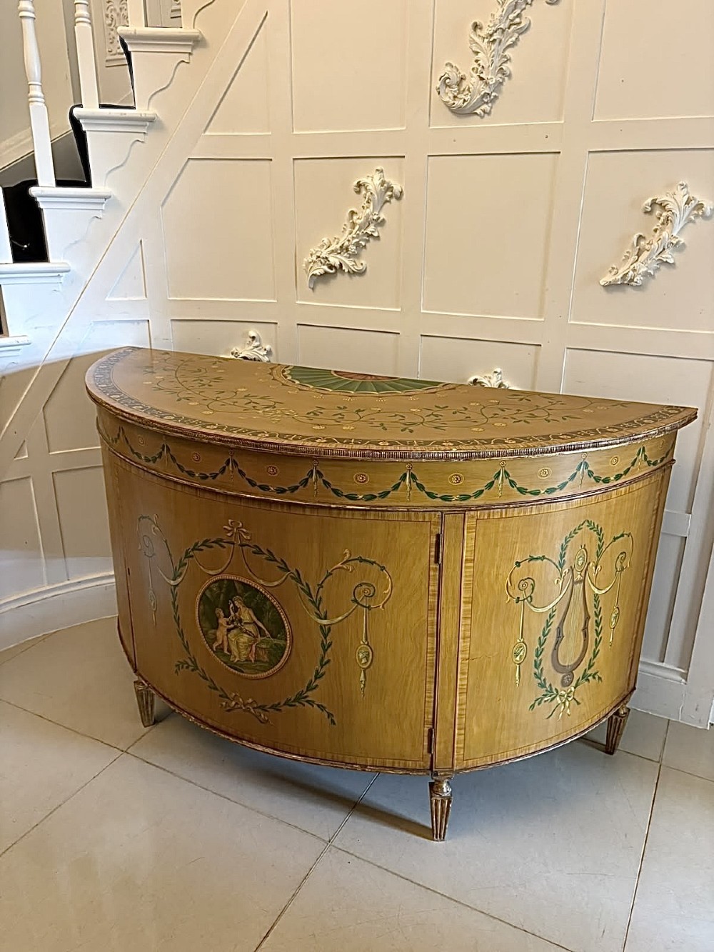 A semicircular satinwood demi-lune cabinet featuring hand-painted neoclassical leaf swags, a central figural medallion, and short tapered feet. View XIV