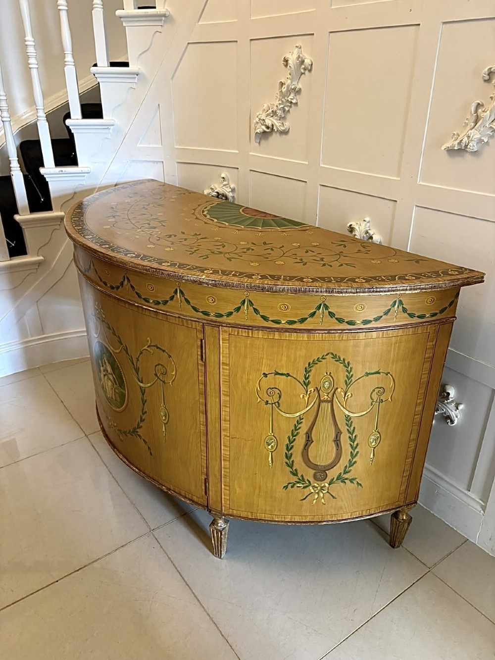 A semicircular satinwood demi-lune cabinet featuring hand-painted neoclassical leaf swags, a central figural medallion, and short tapered feet. View XV