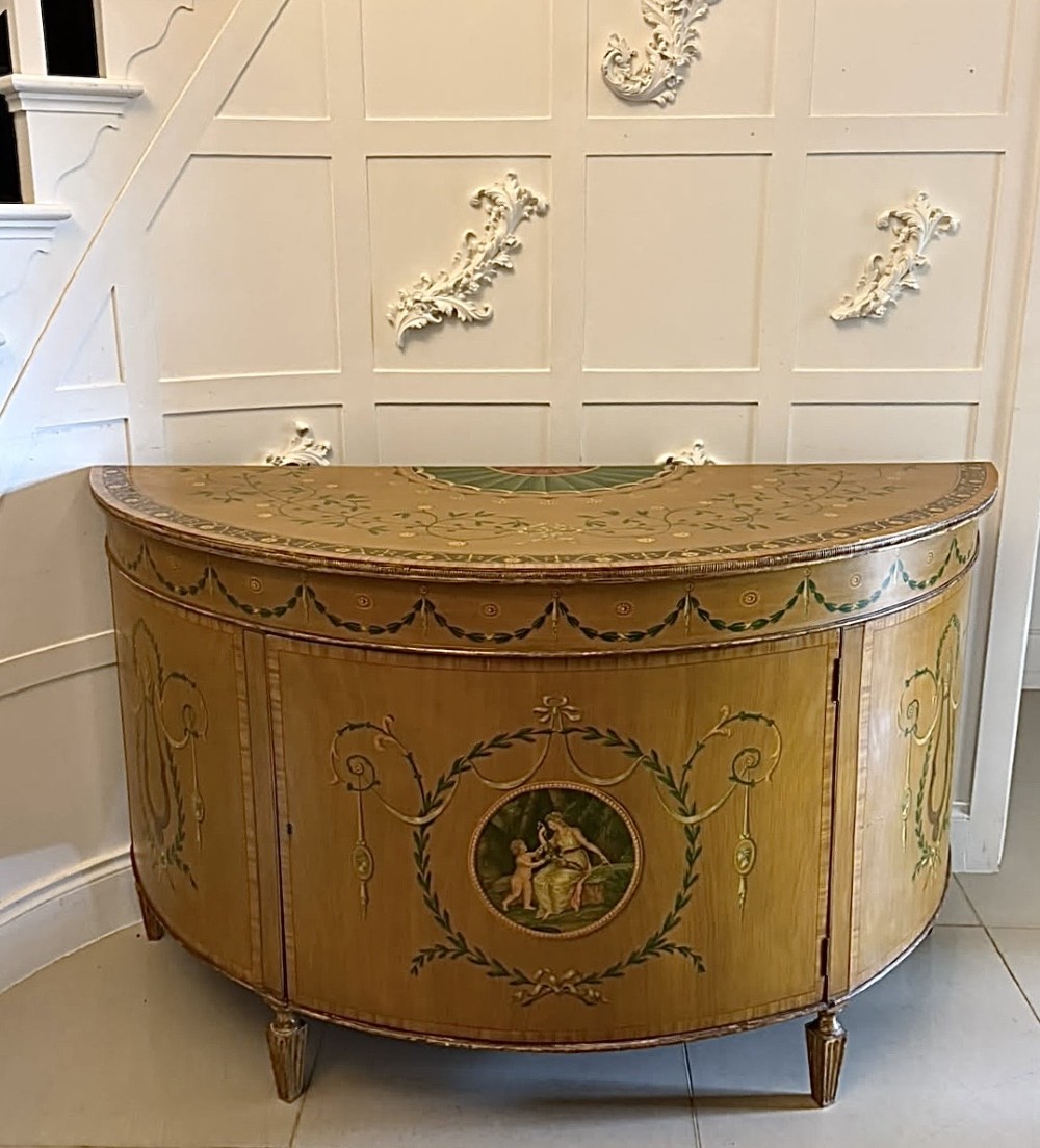 A semicircular satinwood demi-lune cabinet featuring hand-painted neoclassical leaf swags, a central figural medallion, and short tapered feet.