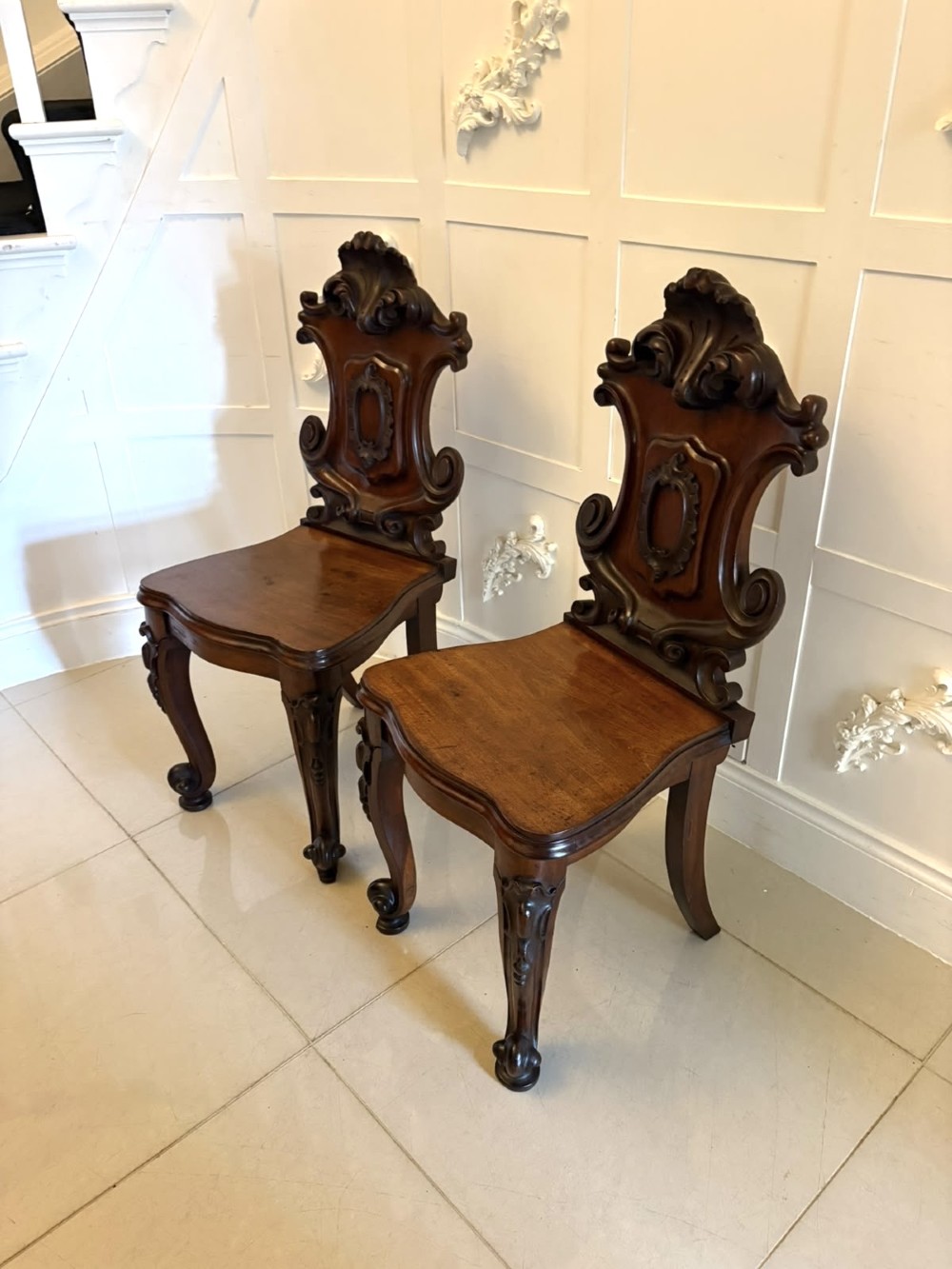 A pair of Victorian carved mahogany hall chairs with shaped timber seats, scrolled cartouche backrests, and carved cabriole legs. View XIV