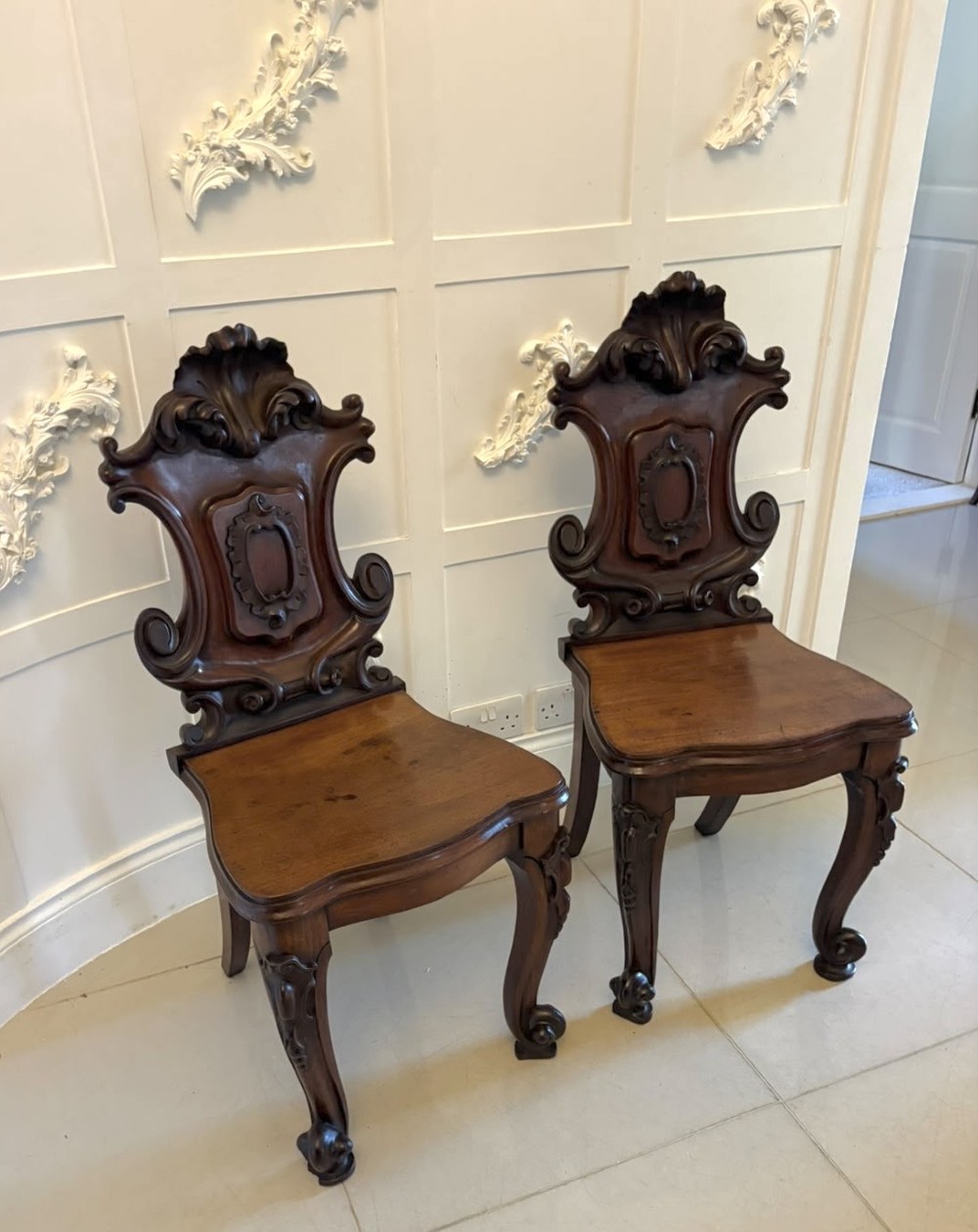 A pair of Victorian carved mahogany hall chairs with shaped timber seats, scrolled cartouche backrests, and carved cabriole legs. View XV