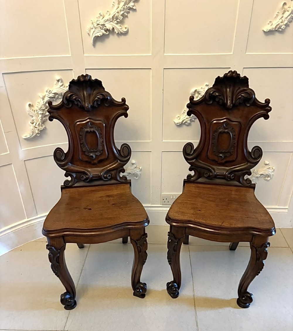 A pair of Victorian carved mahogany hall chairs with shaped timber seats, scrolled cartouche backrests, and carved cabriole legs.